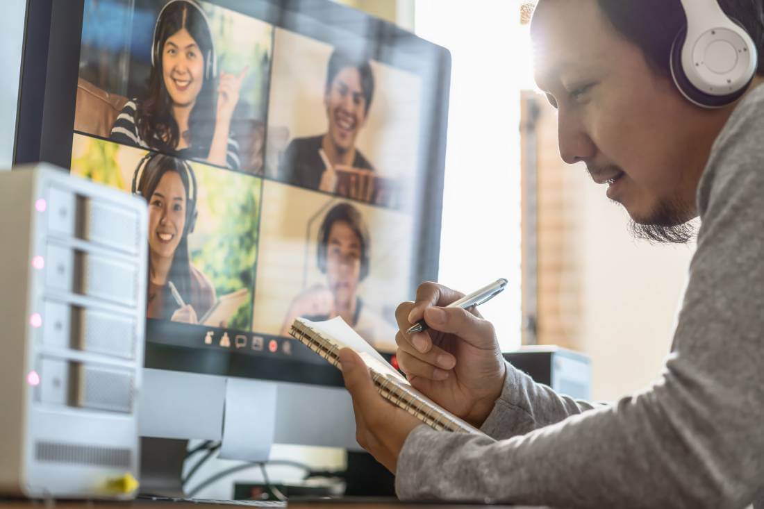 A remote worker takes notes while on his video call with four other colleagues.